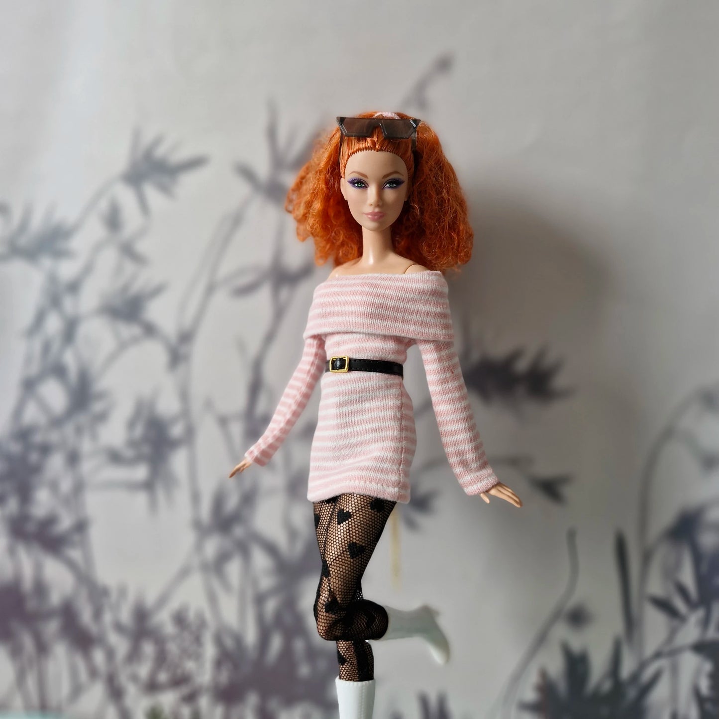 Barbie doll with red hair and a pink sweater standing against a wall with plant shadows.