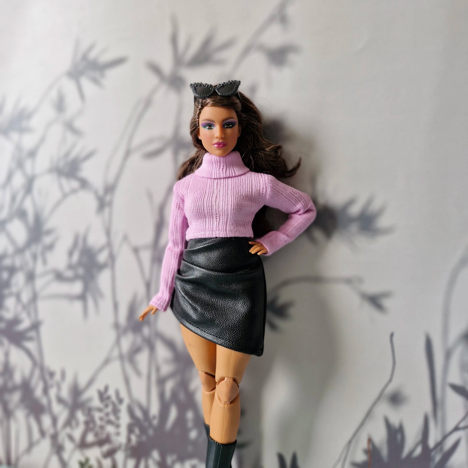 Curvy Barbie wearing a pink turtleneck sweater and black faux leather skirt posing against a soft gray floral background.
