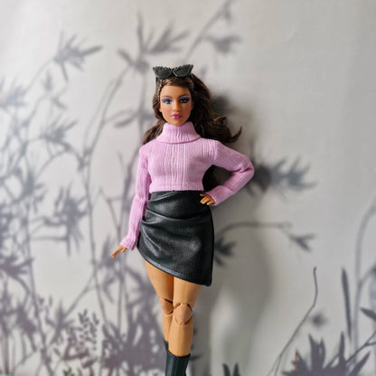 Curvy Barbie wearing a pink turtleneck sweater and black faux leather skirt posing against a soft gray floral background.
