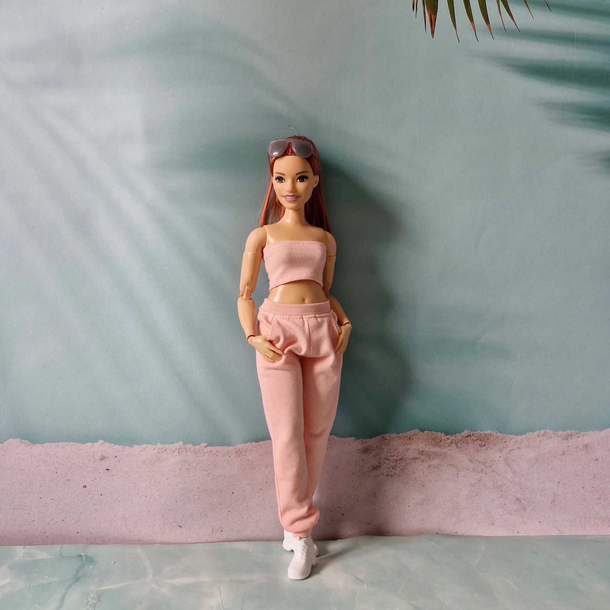 Barbie doll standing in a pink tube top and matching jogger pants against a pastel background.
