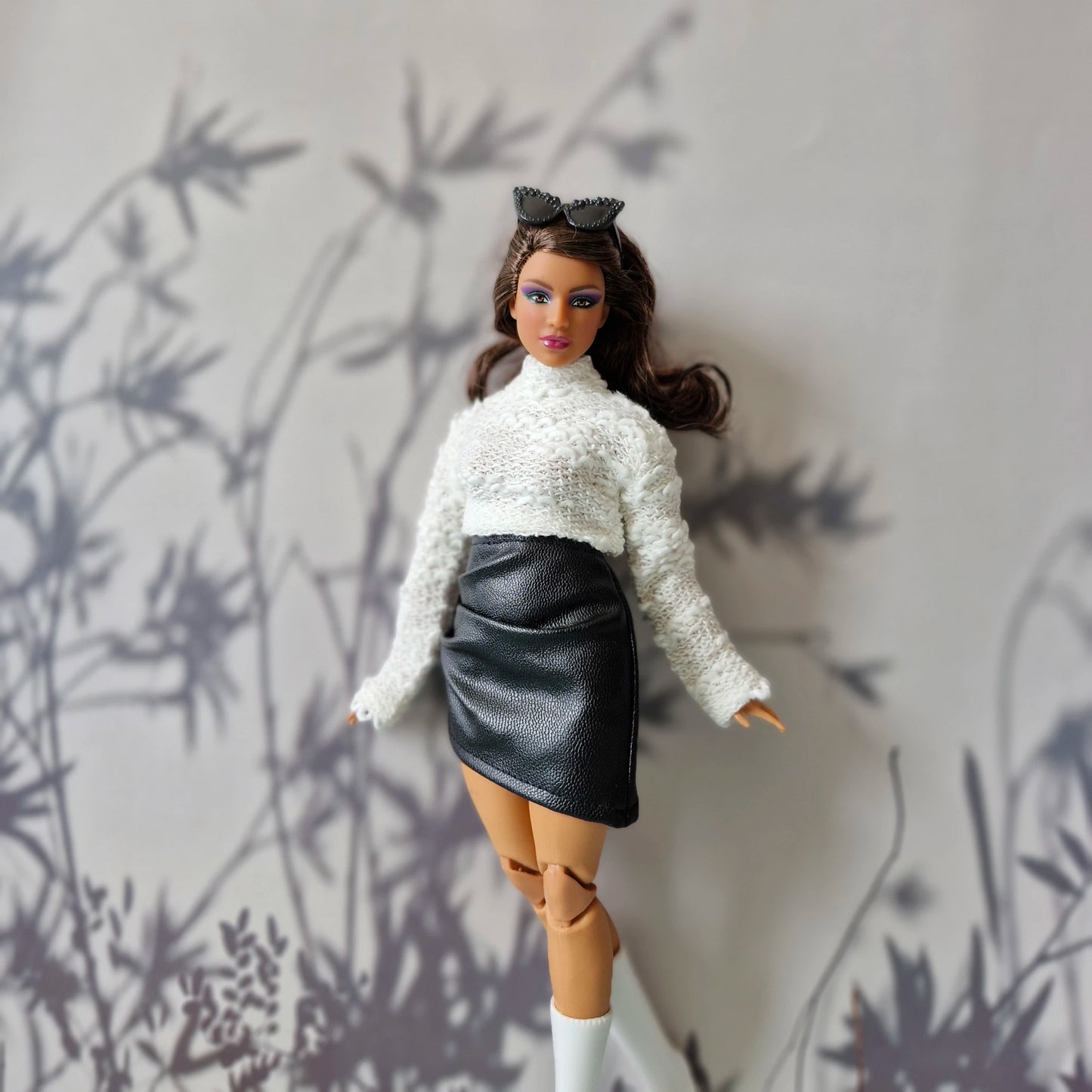 Close-up of Curvy Barbie in white fluffy sweater and black leather skirt posing with soft floral background shadows.
