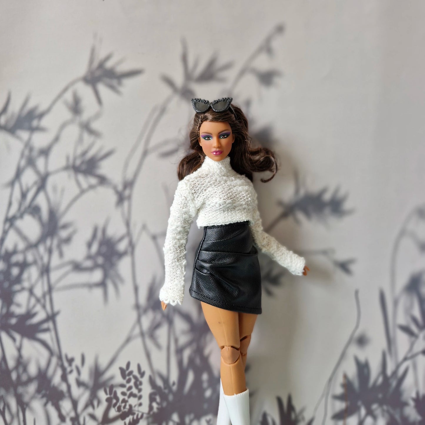 Curvy Barbie in white high-neck sweater and black faux leather skirt standing confidently in front of shadowed floral backdrop.
