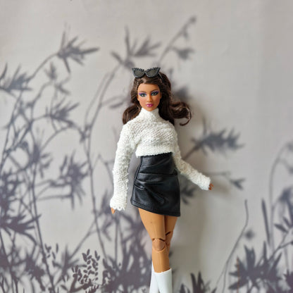 Curvy Barbie in white high-neck sweater and black faux leather skirt standing confidently in front of shadowed floral backdrop.
