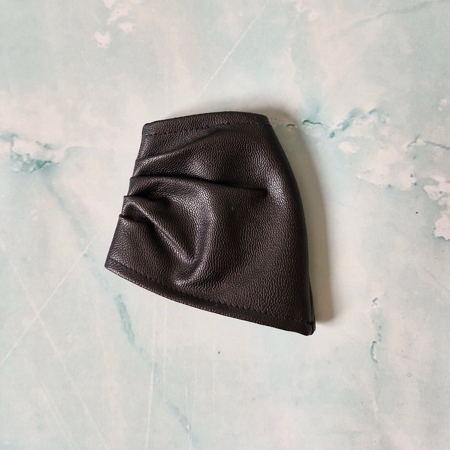 Black faux leather asymmetrical Barbie skirt laid flat on a marble-textured surface.
