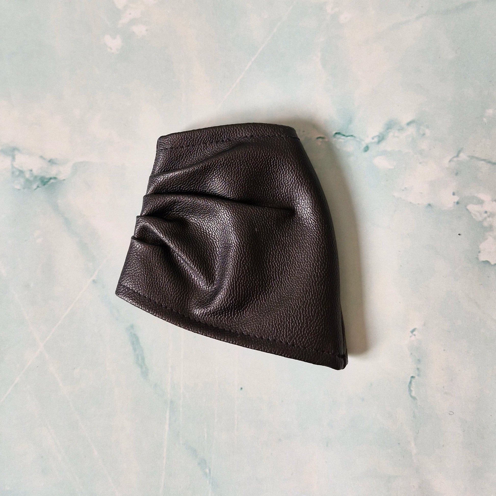 Black faux leather asymmetrical Barbie skirt laid flat on a marble-textured surface.

