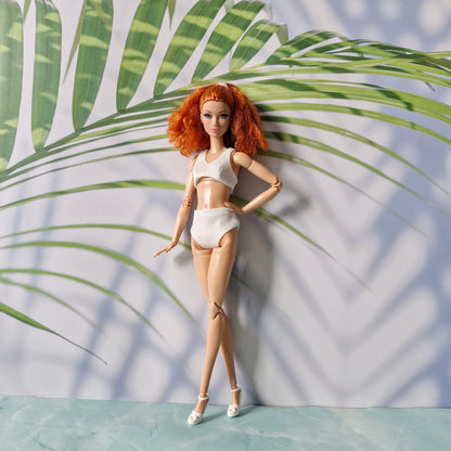 Red-haired Barbie doll wearing a white bikini set posing against pastel tropical leaf background.
