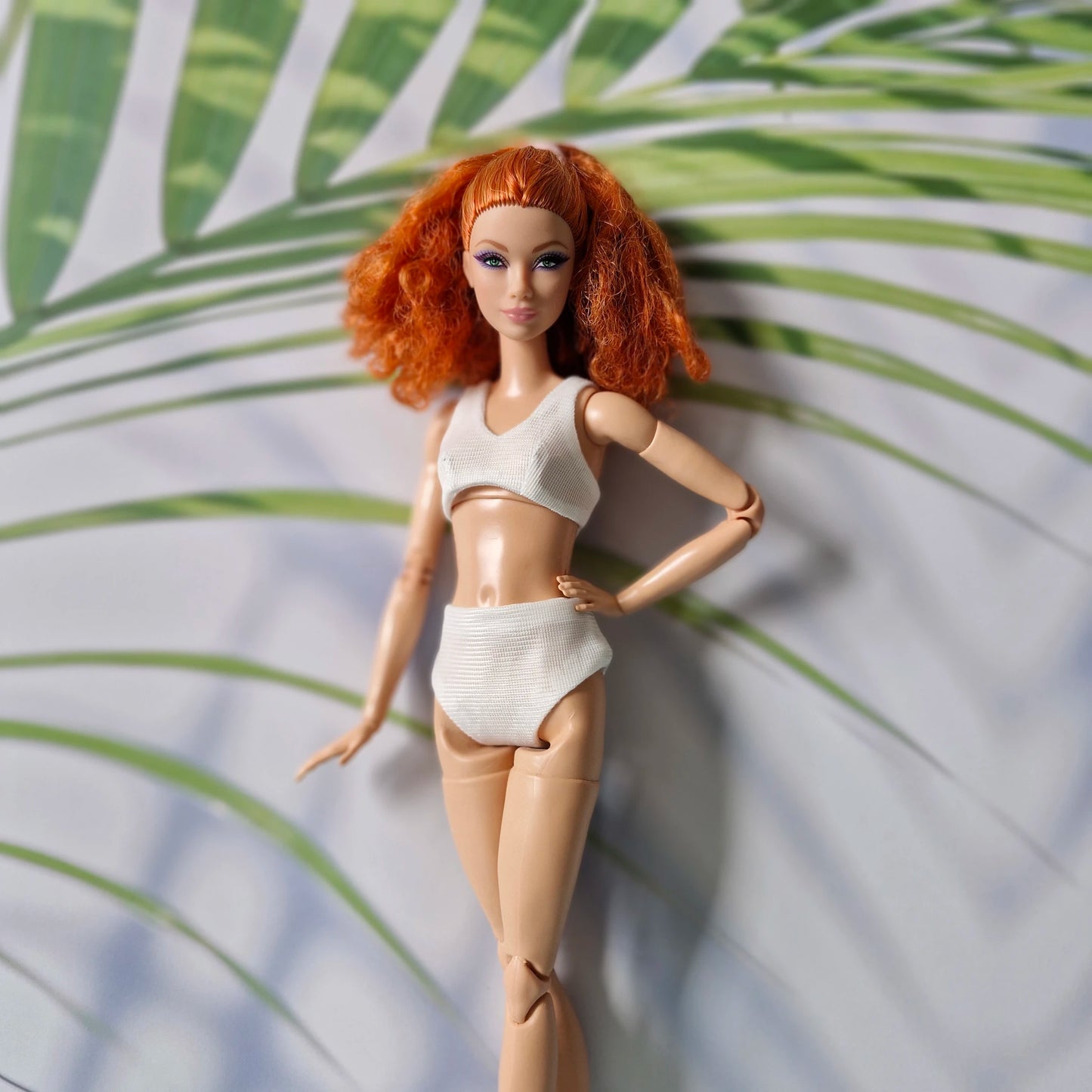 Close-up of red-haired Barbie doll in a white ribbed bikini with tropical leaf background.
