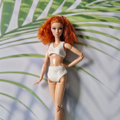 Close-up of red-haired Barbie doll in a white ribbed bikini with tropical leaf background.
