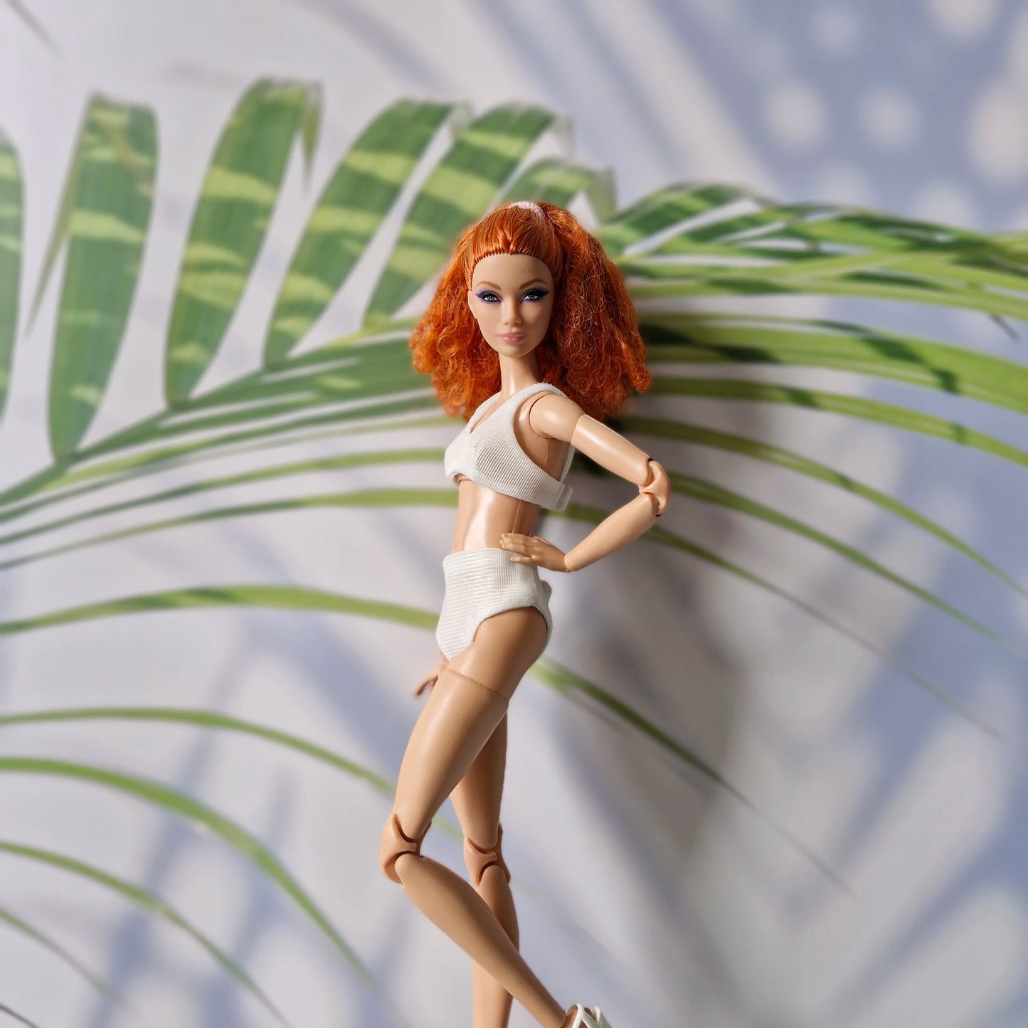 Barbie doll with red curly hair wearing a white ribbed bikini set posing in front of tropical leaves.
