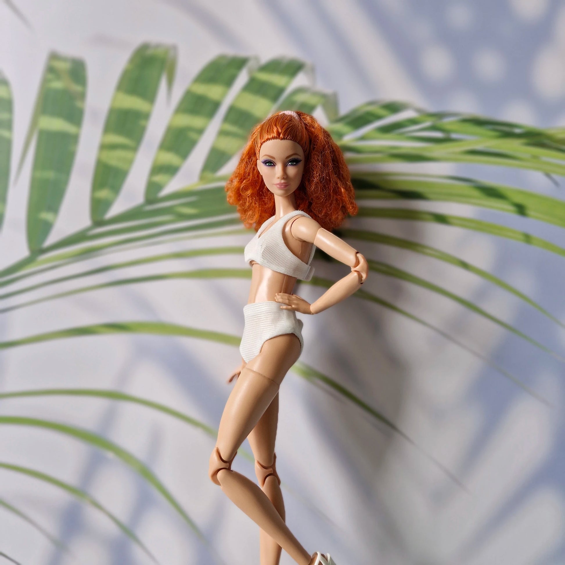 Barbie doll with red curly hair wearing a white ribbed bikini set posing in front of tropical leaves.
