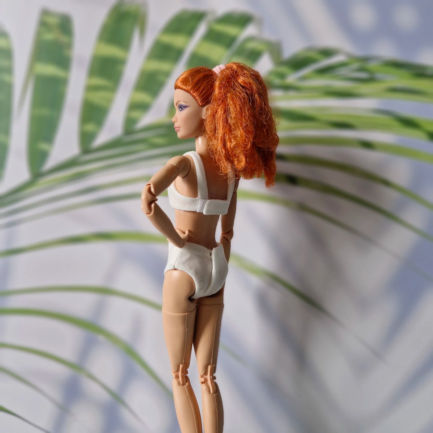 Back view of red-haired Barbie doll wearing a white ribbed bikini set in a tropical leaf background.
