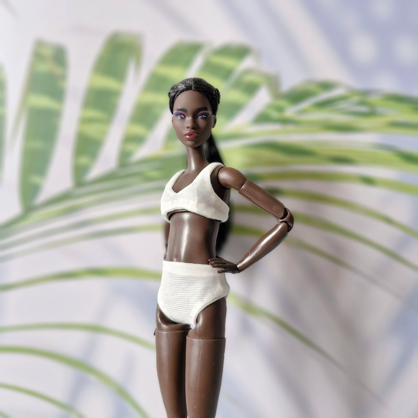 Dark-skinned Barbie doll wearing a white ribbed bikini top and high-waisted bottoms standing in front of palm leaves.
