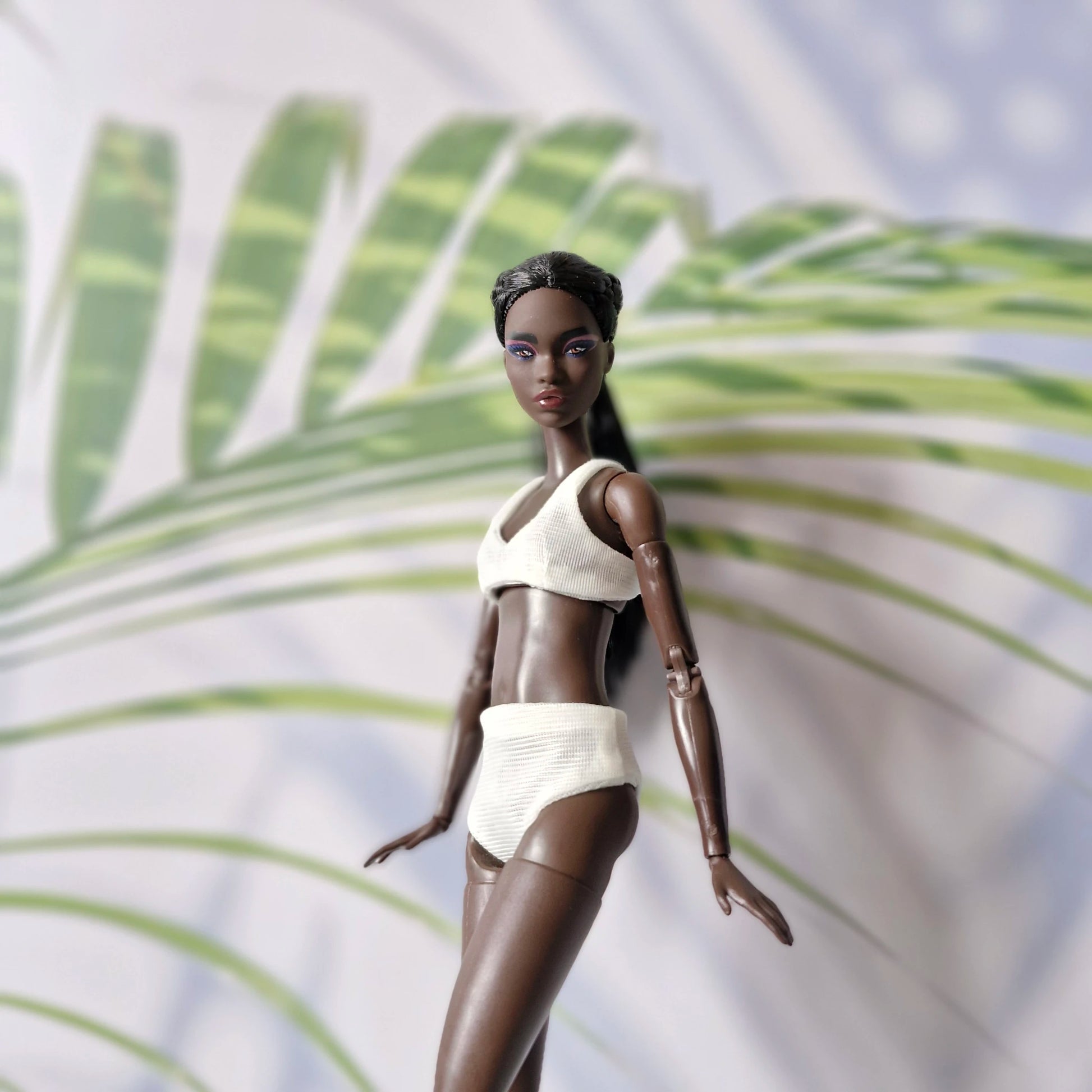 Side-pose dark-skinned Barbie doll in a white ribbed bikini against a leaf shadow background.
