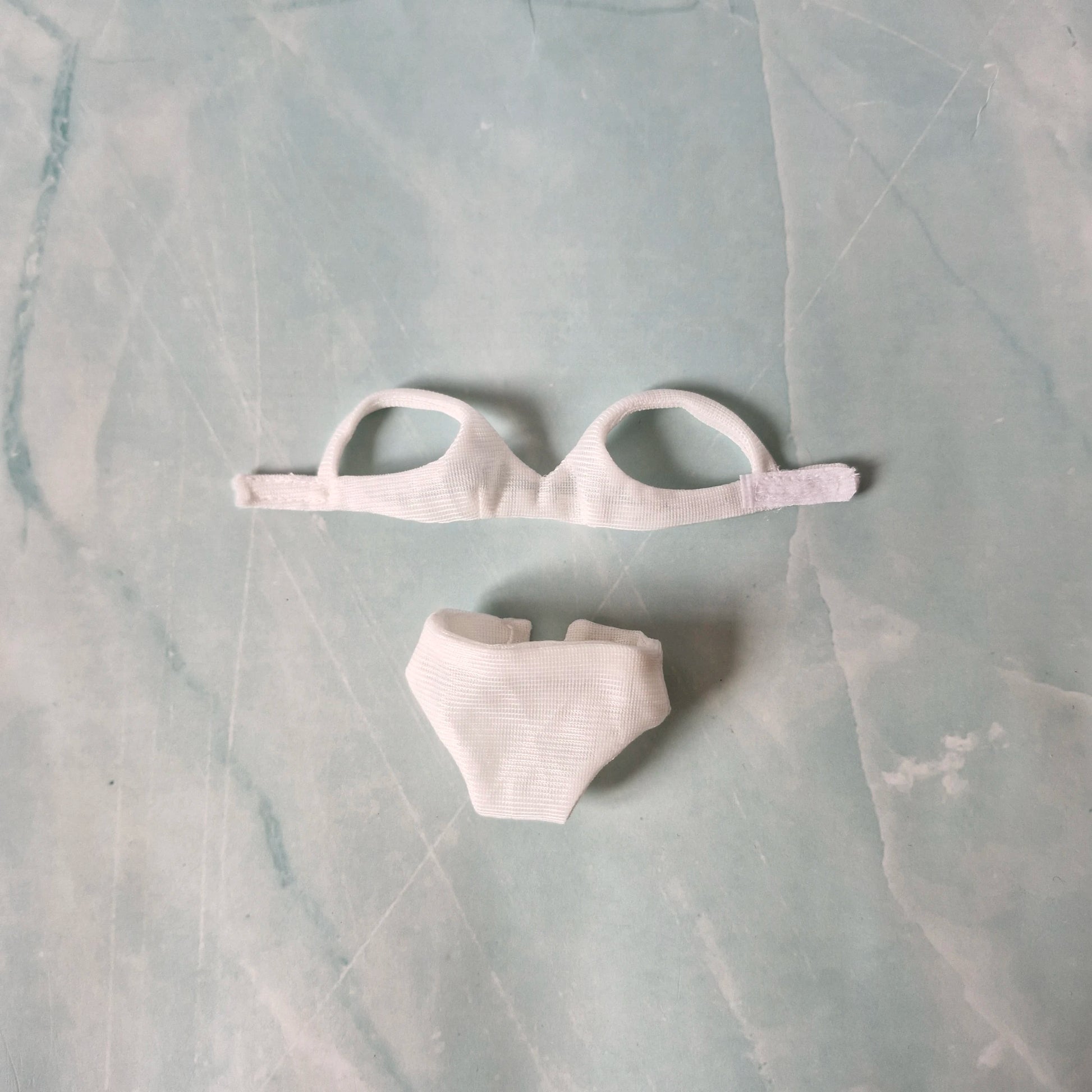 Flat lay of white ribbed bikini top and high-waisted bottoms for Barbie dolls on a pastel background.
