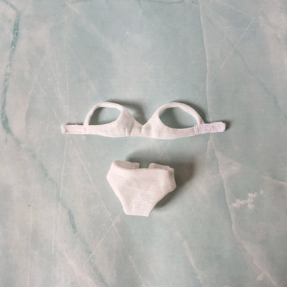 Flat lay of white ribbed bikini top and high-waisted bottoms for Barbie dolls on a pastel background.
