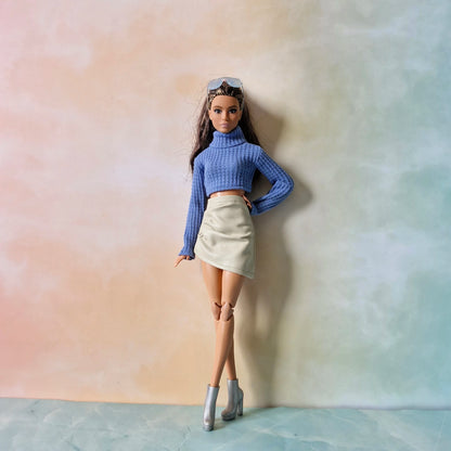 Barbie doll wearing blue cropped turtleneck sweater and cream faux-leather skirt posing against soft gradient background.
