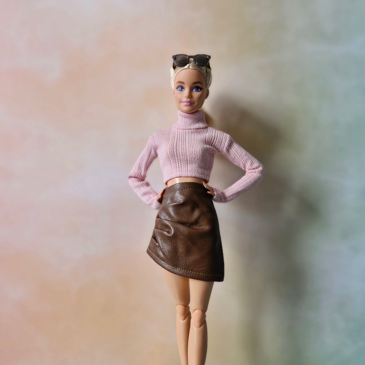 Blonde Barbie in pink sweater and brown faux-leather skirt standing in pastel backdrop.
