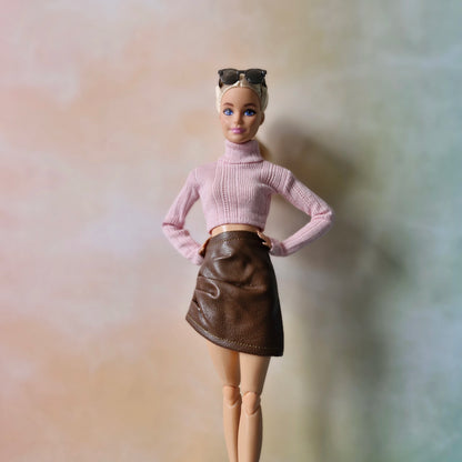 Blonde Barbie in pink sweater and brown faux-leather skirt standing in pastel backdrop.
