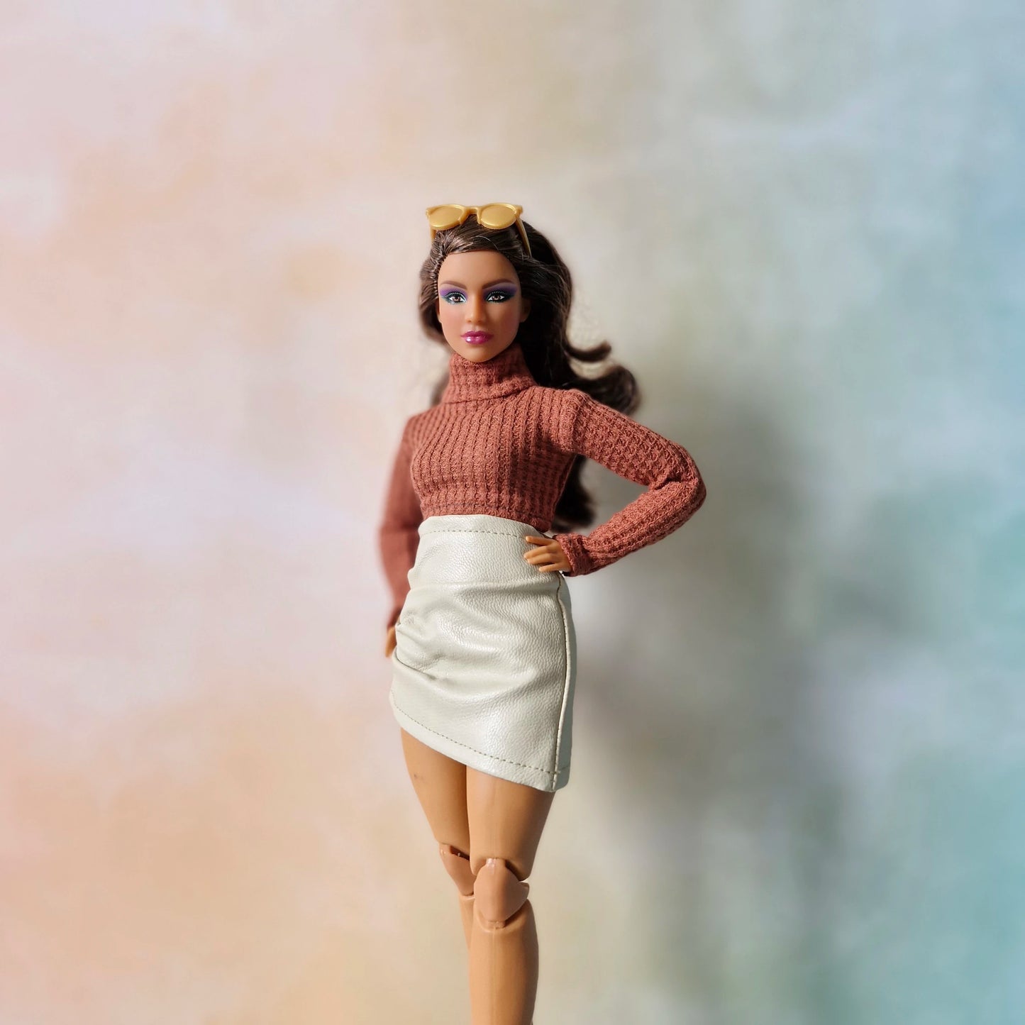 Tan-skin Barbie wearing rust turtleneck sweater and cream faux-leather skirt posing against gradient background.
