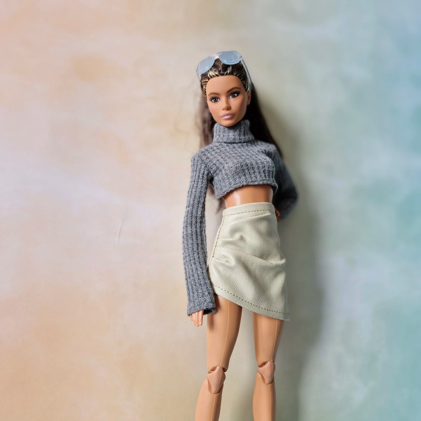 Barbie wearing grey cropped turtleneck sweater and cream faux-leather skirt against pastel gradient background.
