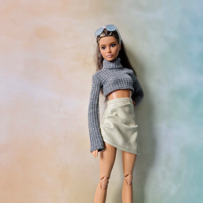 Barbie wearing grey cropped turtleneck sweater and cream faux-leather skirt against pastel gradient background.
