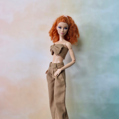Barbie with red curly hair wearing beige brown bow top and wide-leg pants on pastel background.
