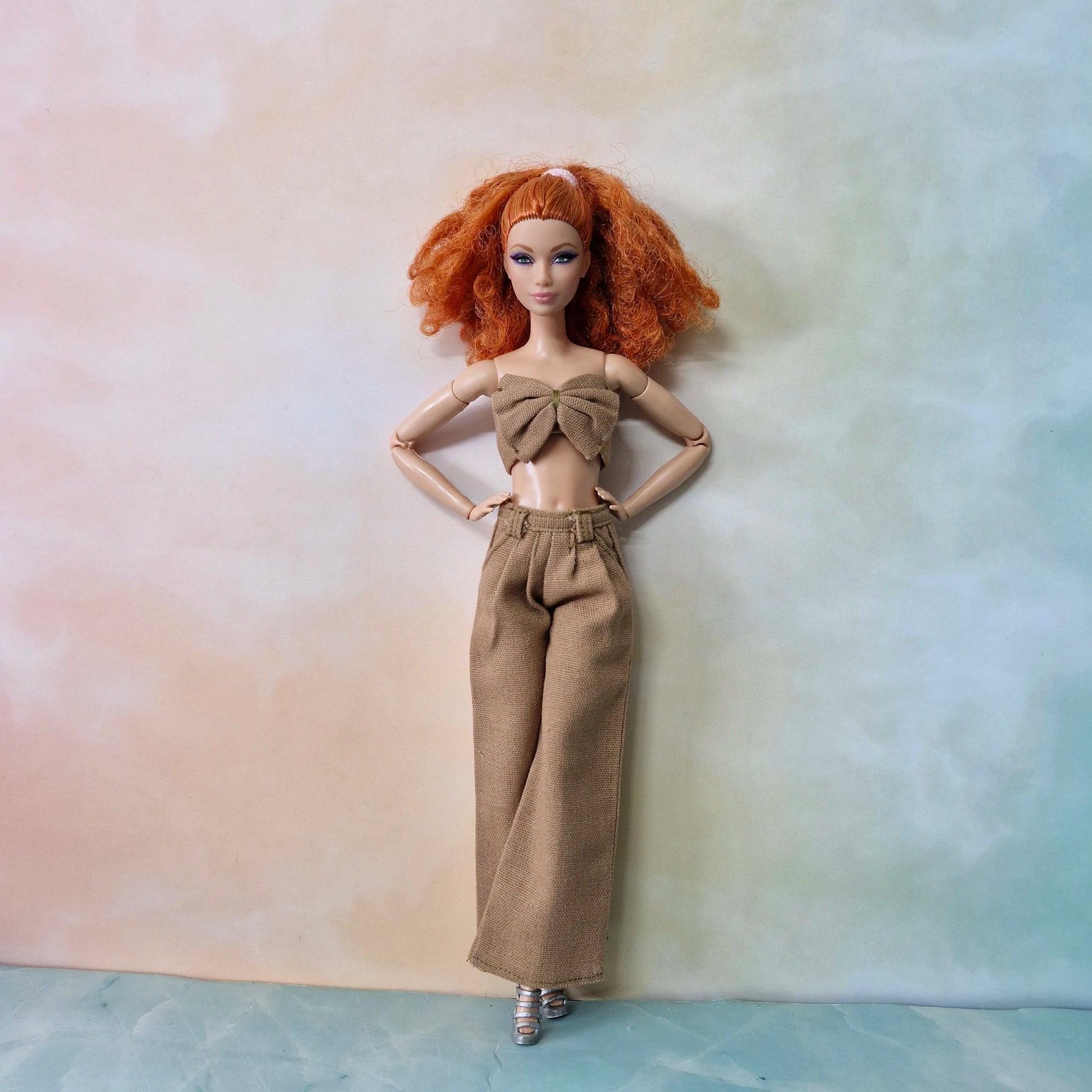 Full-body Barbie in beige brown bow top and wide-leg trousers posing in soft pastel scene.
