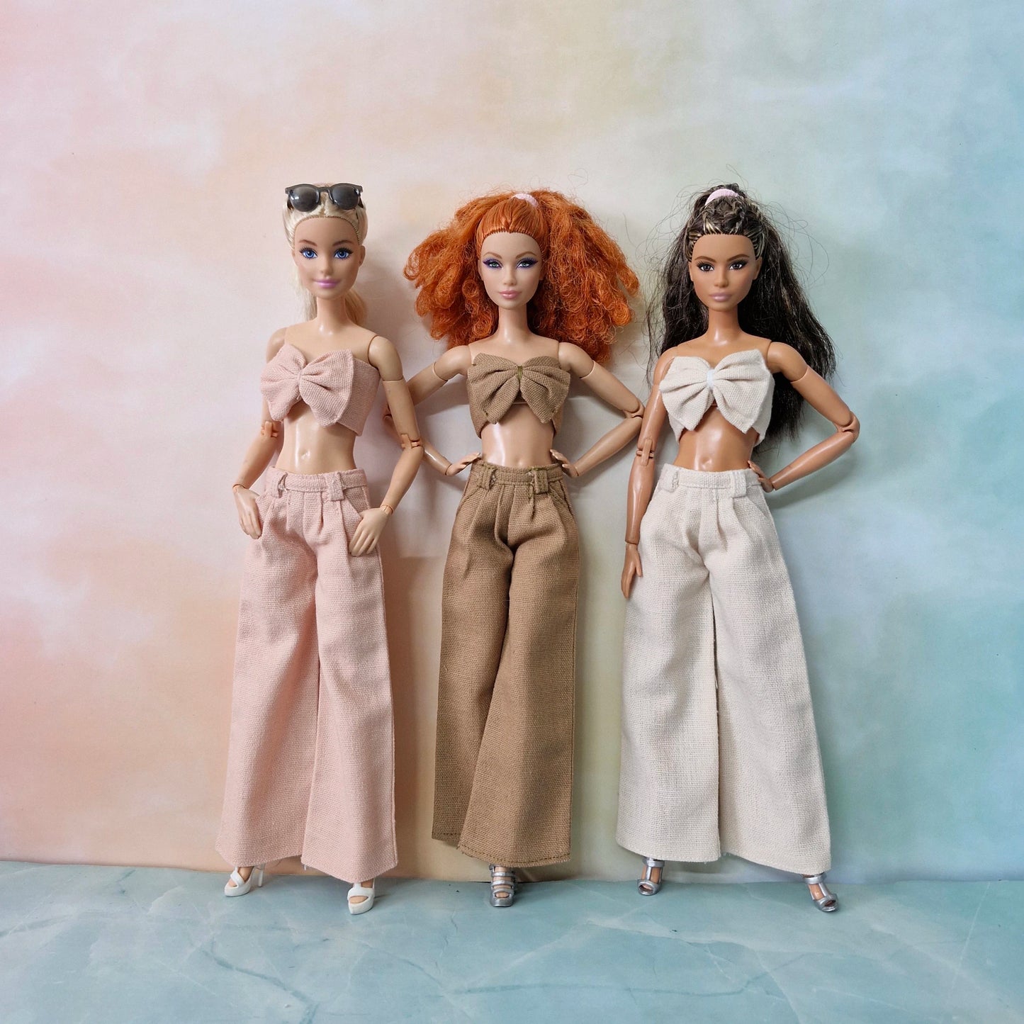 Three Barbie dolls wearing bow top and wide-leg pants in blush pink, beige brown, and cream tones.
