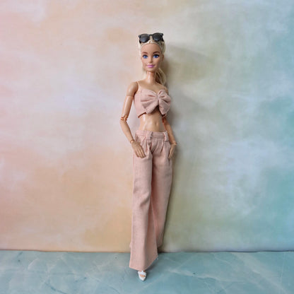 Full-body Barbie in blush pink bow top and wide-leg trousers posing against gradient pastel backdrop.

