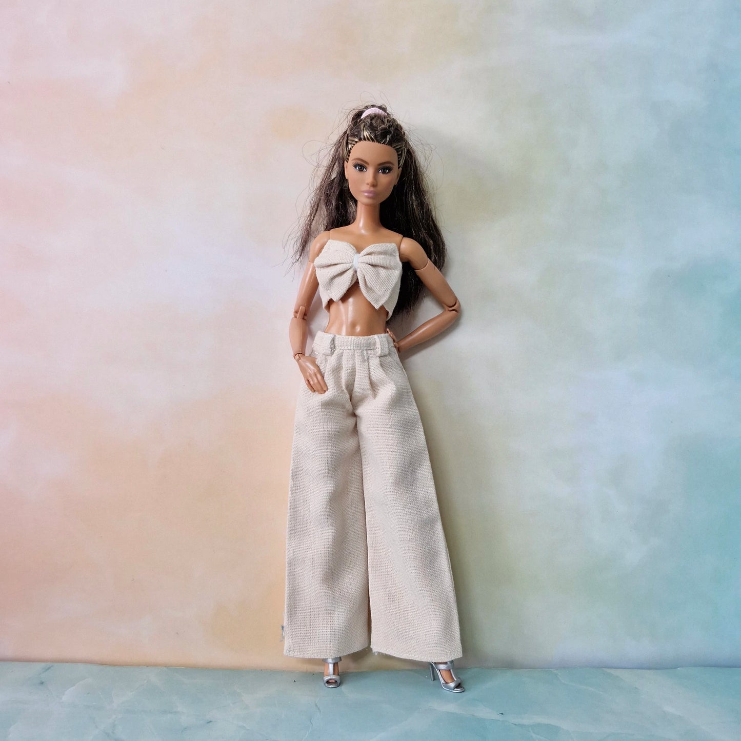 Brunette Barbie wearing cream bow top and matching cream wide-leg pants on soft pastel background.

