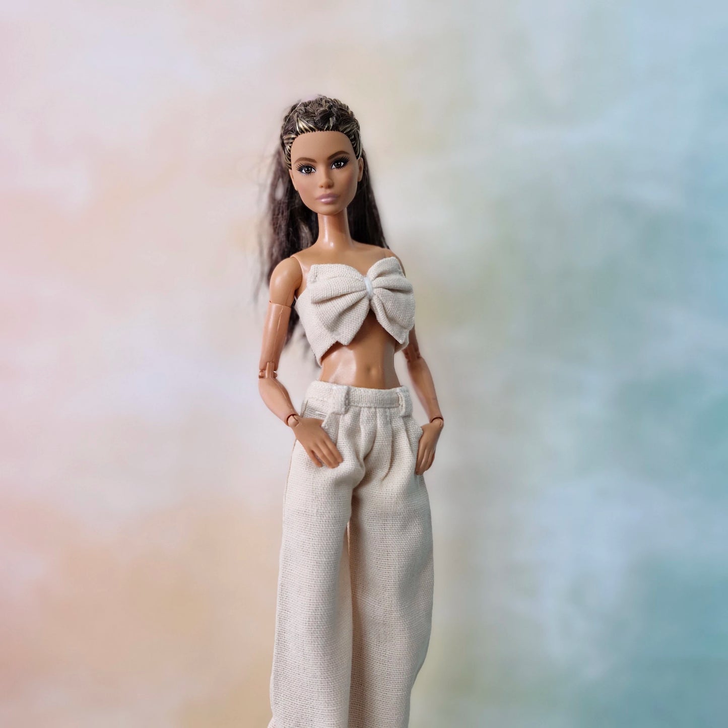 Close-up of Barbie in cream bow top and wide-leg pants posing against pastel gradient.
