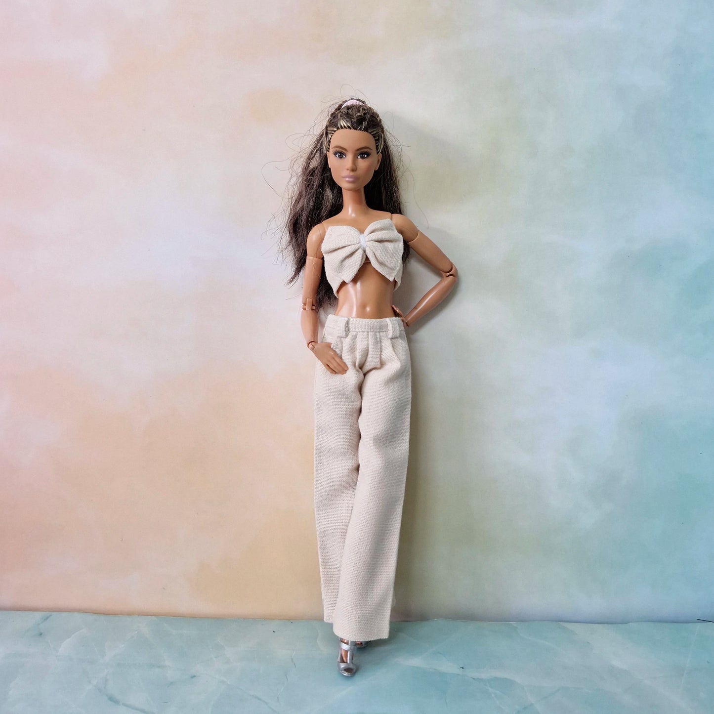 Barbie model wearing cream bow top and wide-leg pants standing against pastel backdrop.
