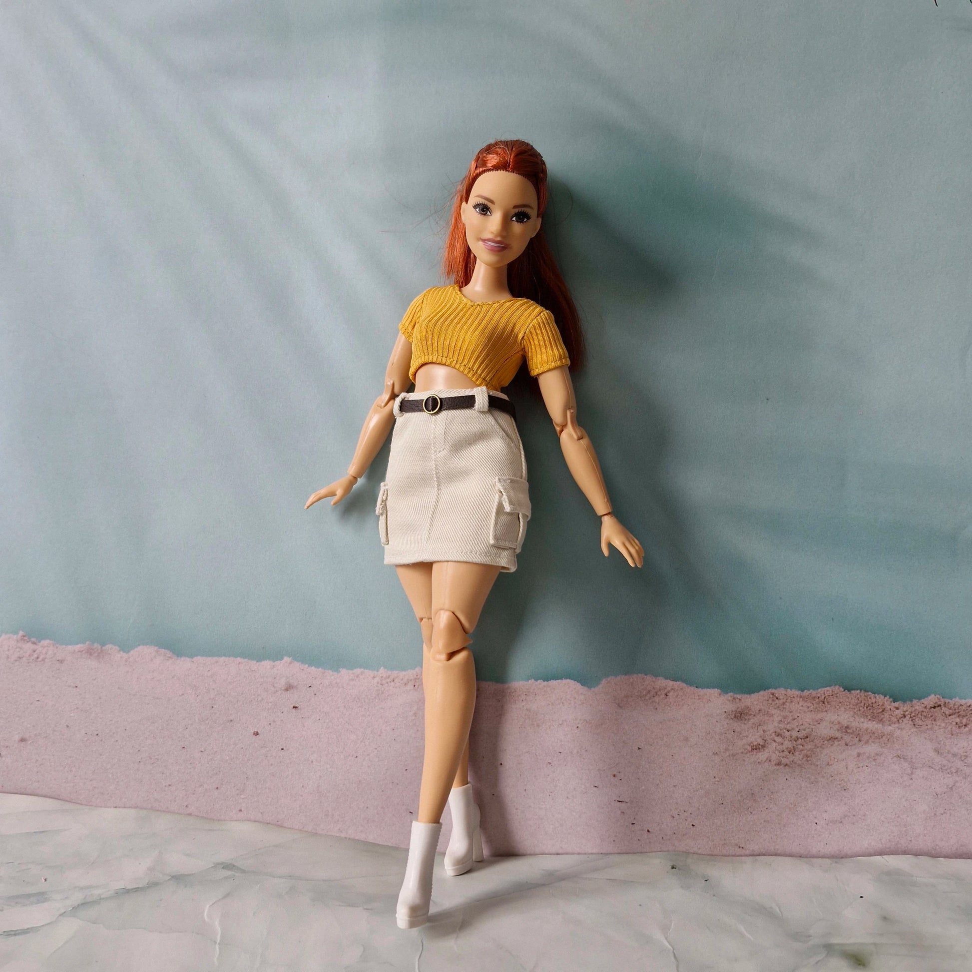 Fashion doll modeling casual outfit with yellow crop top and beige skirt against blue background
