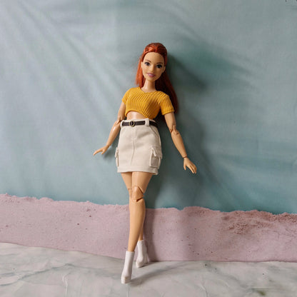 Fashion doll modeling casual outfit with yellow crop top and beige skirt against blue background