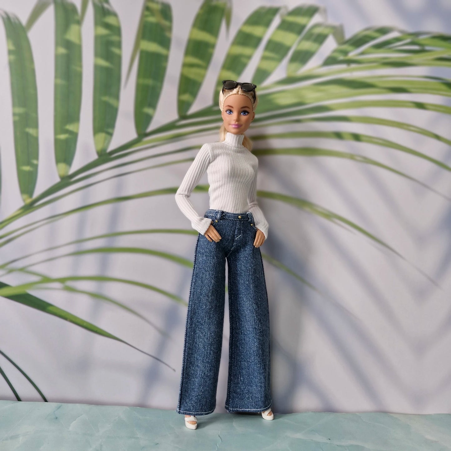 Blonde Barbie doll wearing a white ribbed turtleneck and dark blue wide leg jeans standing against a tropical background