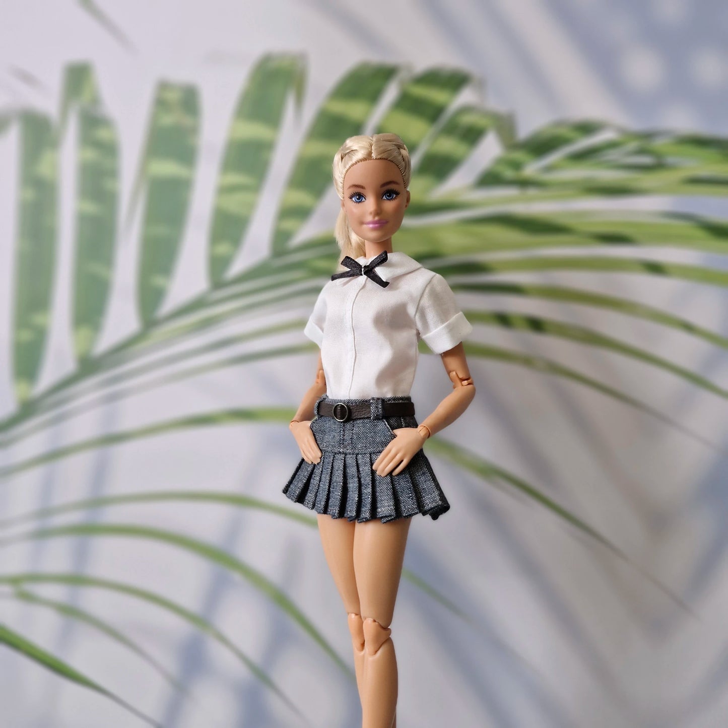 Barbie doll in a white shirt and black pleated skirt standing against a leafy background