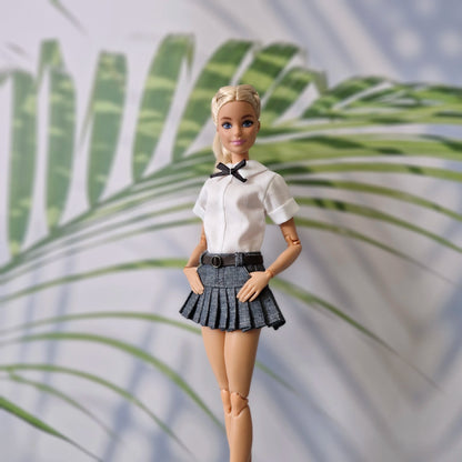 Barbie doll in a white shirt and black pleated skirt standing against a leafy background