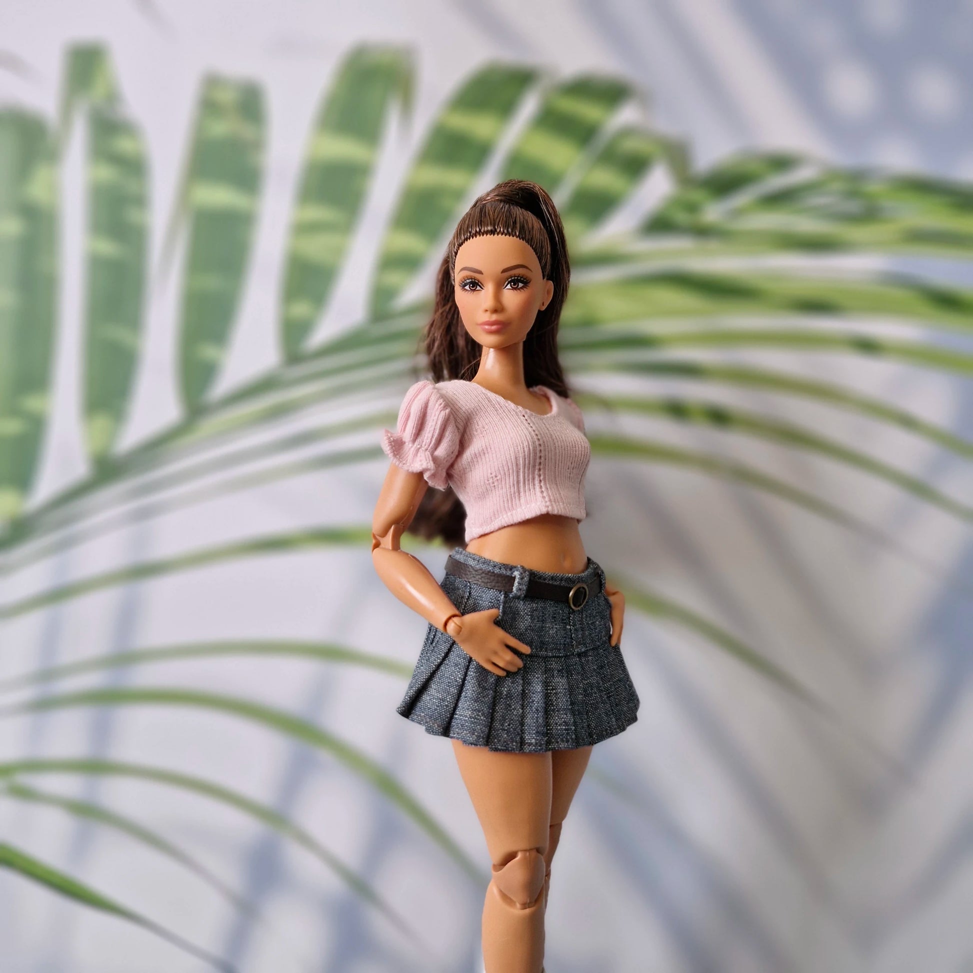 Barbie doll in a pink top and plaid skirt standing against a palm leaf background