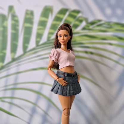 Barbie doll in a pink top and plaid skirt standing against a palm leaf background