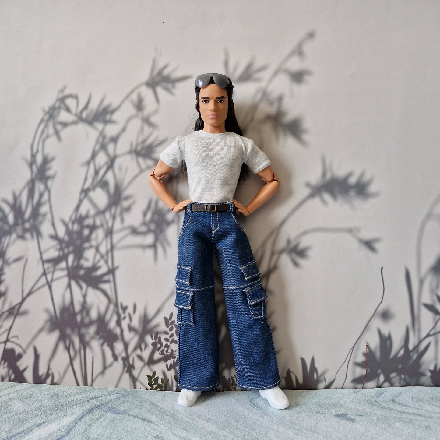 Toy figure wearing a white shirt and blue jeans standing against a wall with plant shadows.