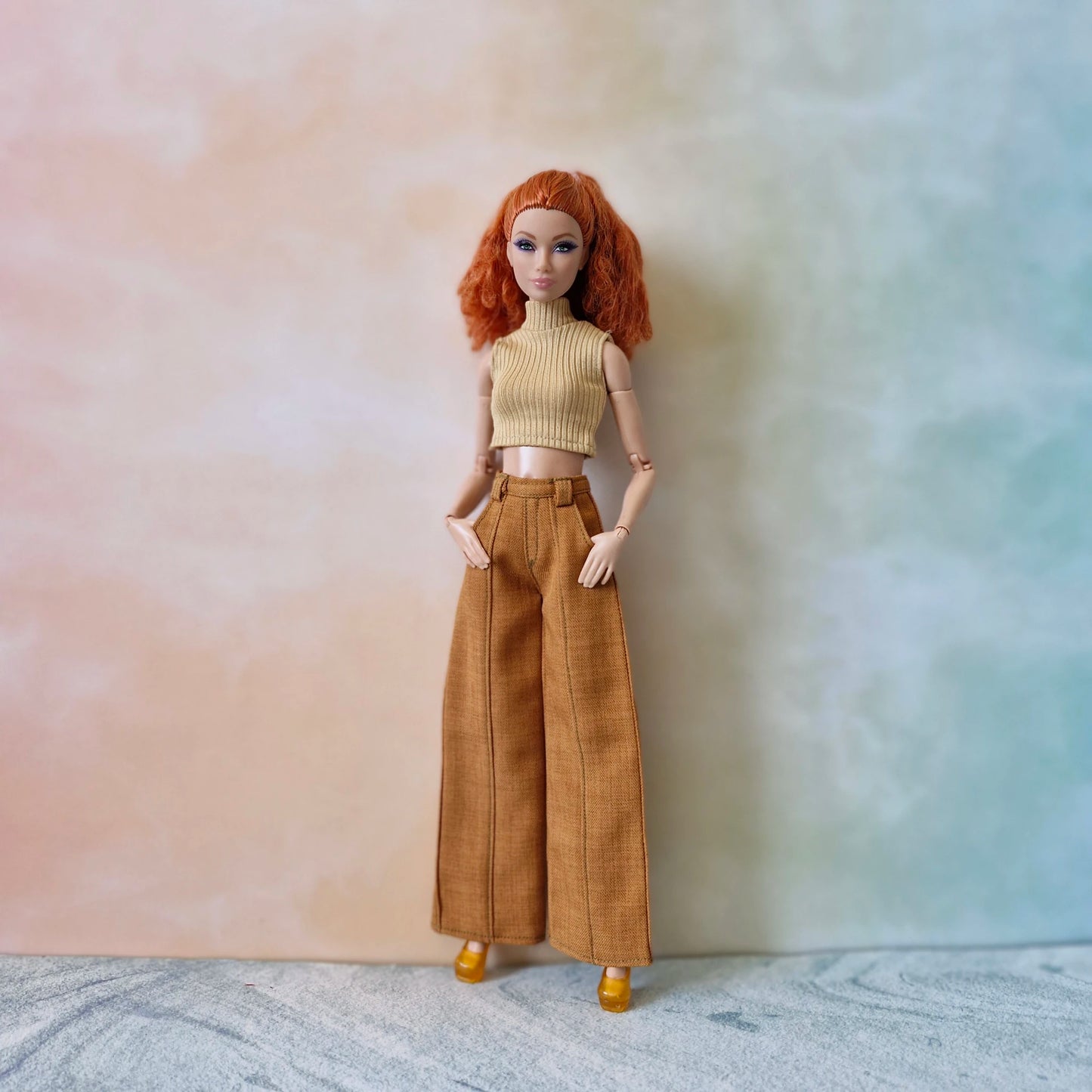 Doll wearing a beige top and brown pants against a gradient background