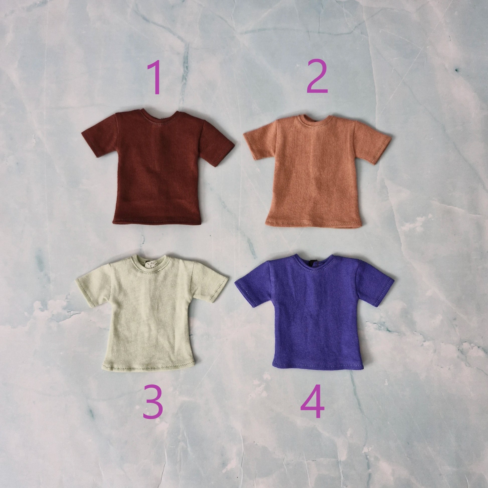 A flat lay image displaying multiple folded Ken doll t-shirts in blue, grey, and cream colors