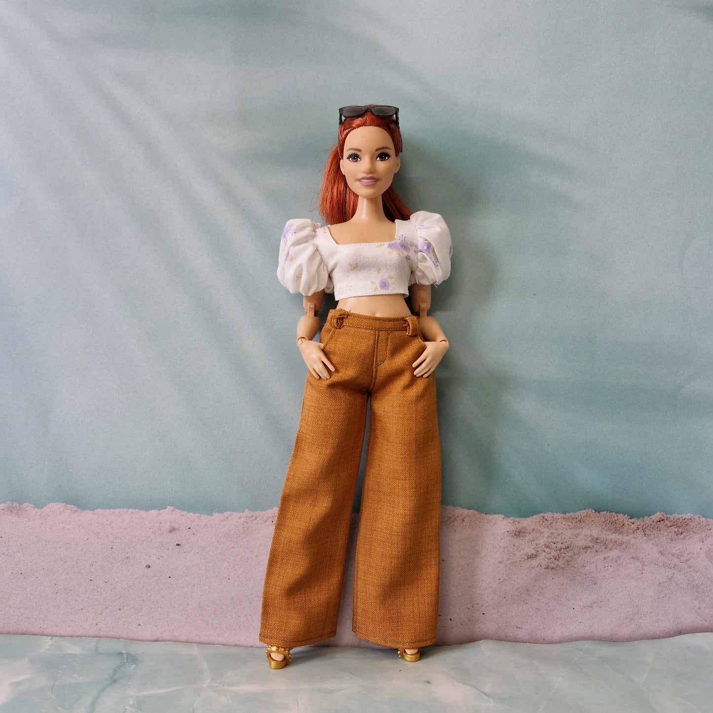 Curvy Barbie doll wearing a white crop top with purple floral print and brown wide leg pants, posing against a beach backdrop.