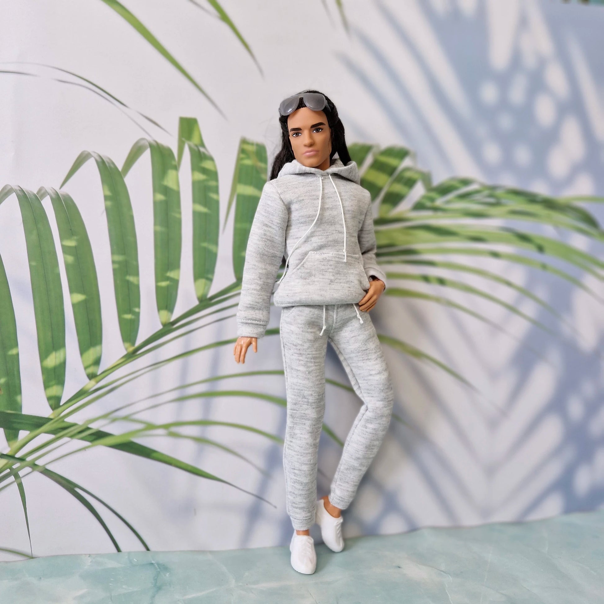 Ken doll wearing a trendy grey activewear set including a pullover hoodie and sweatpants, posing with one leg bent.