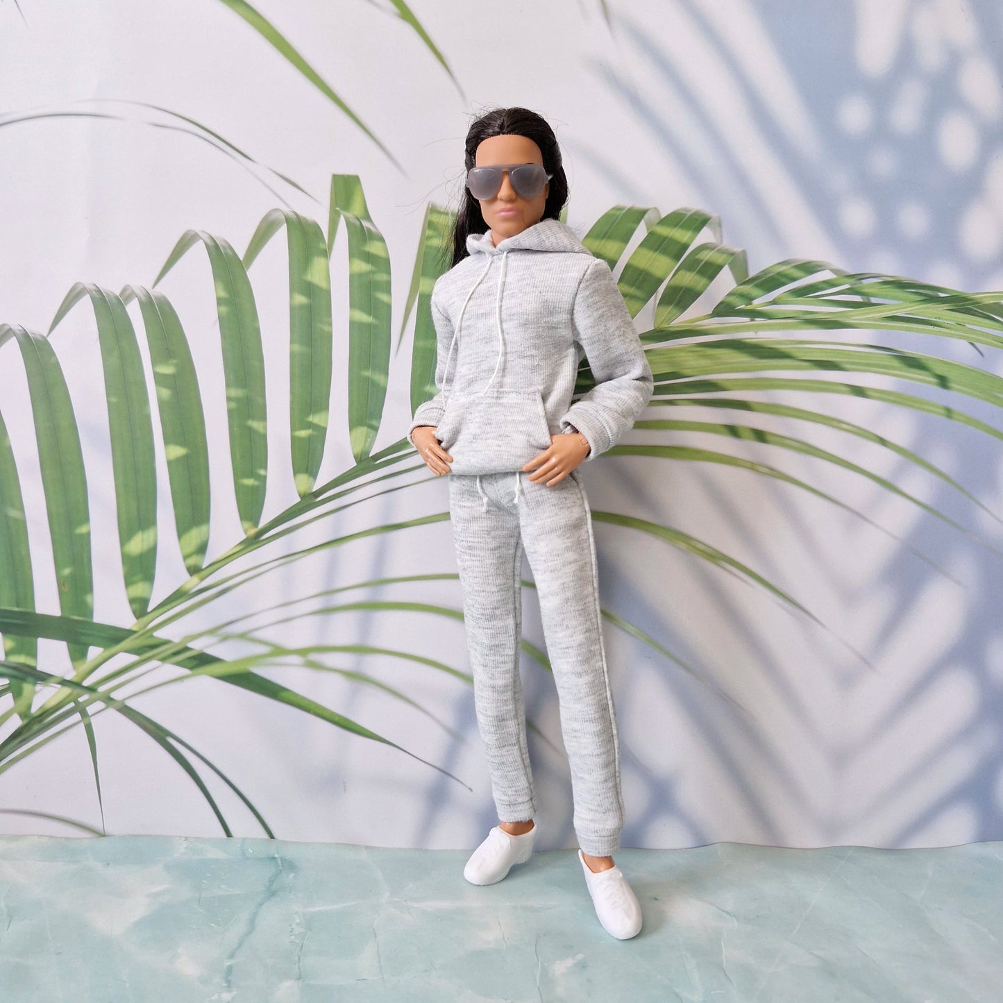 Ken doll wearing a casual grey hoodie and matching sweatpants, posing with hands in his pocket against a tropical leaf background.