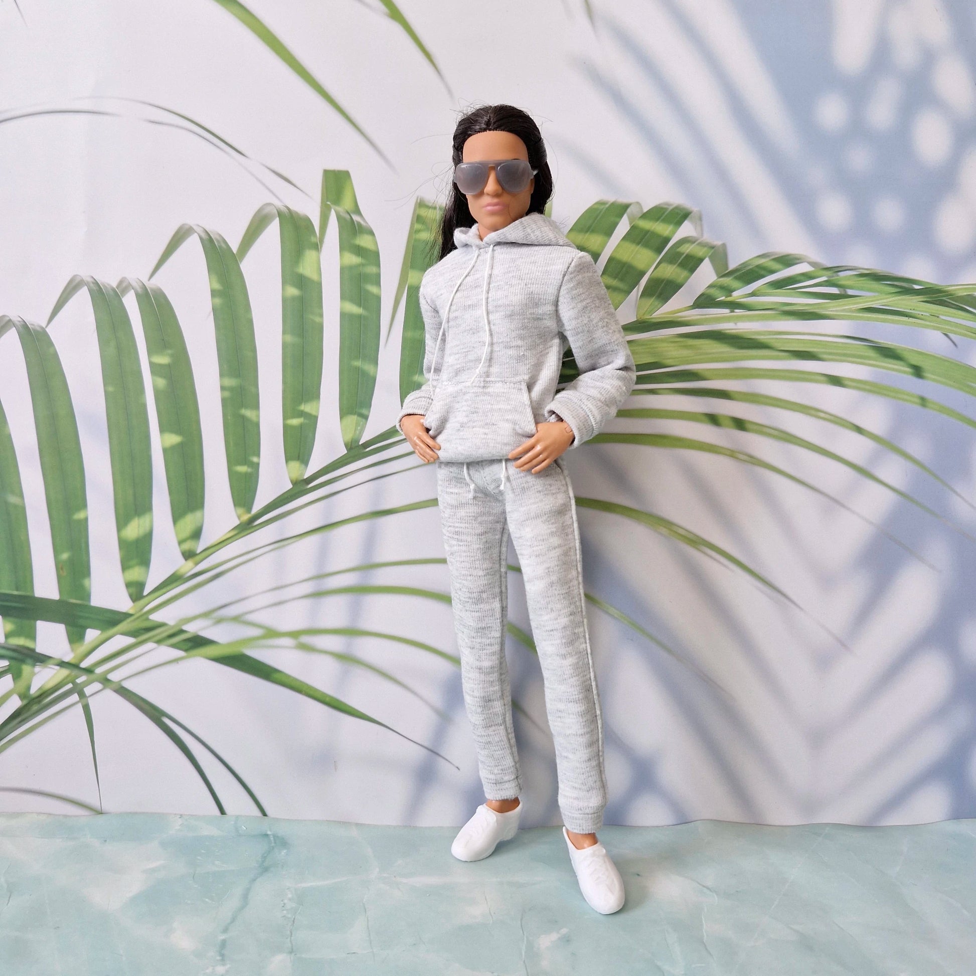 Ken doll wearing a casual grey hoodie and matching sweatpants, posing with hands in his pocket against a tropical leaf background.