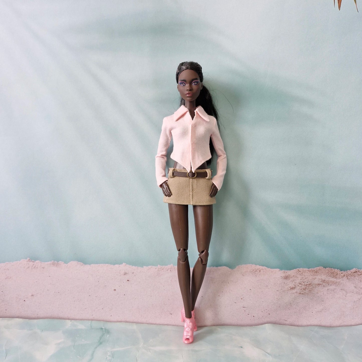 Dark-skinned Barbie doll posing in a light pink pointed hem shirt and beige belted mini skirt.