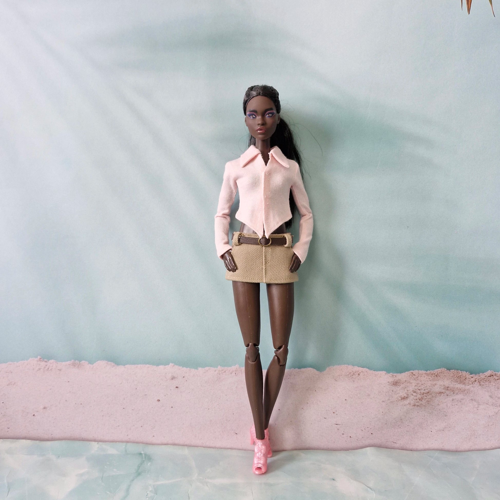 Dark-skinned Barbie doll posing in a light pink pointed hem shirt and beige belted mini skirt.