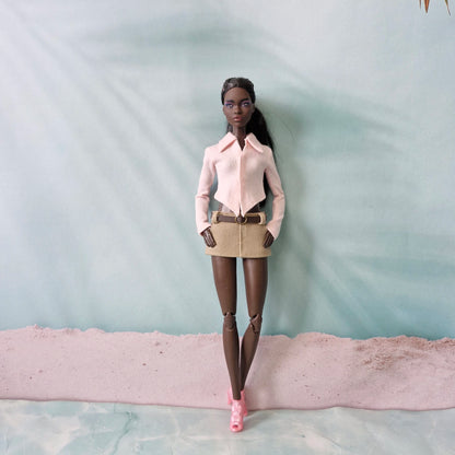 Dark-skinned Barbie doll posing in a light pink pointed hem shirt and beige belted mini skirt.