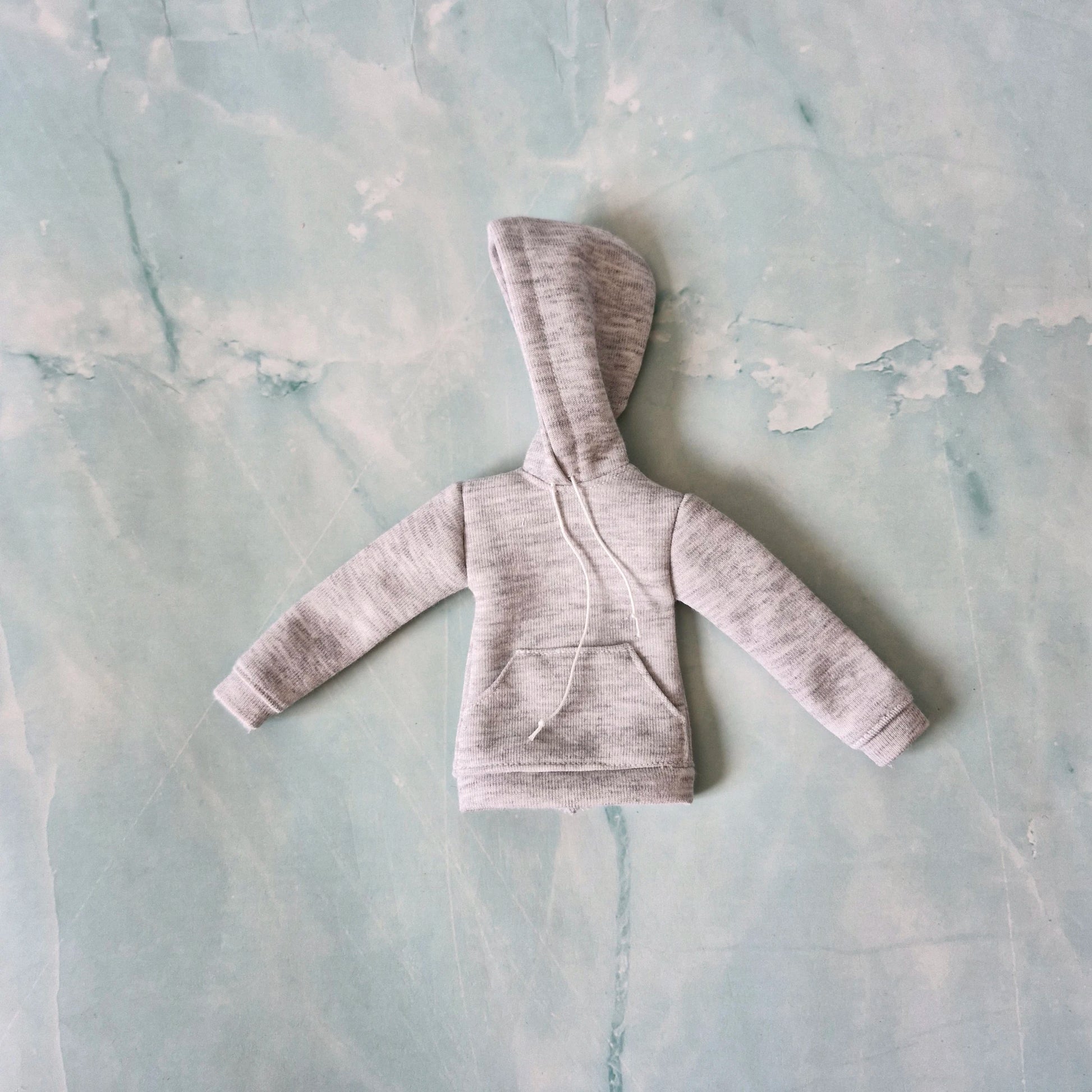 Flat lay of a grey miniature doll-sized hoodie featuring a front kangaroo pocket and white hood drawstrings.